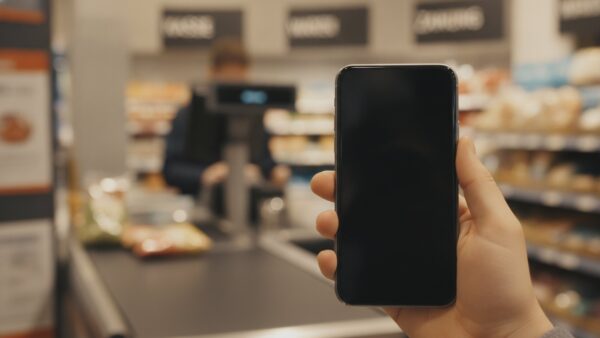 payback login bild 4 Hand hält ein Smartphone mit dem Daumen auf dem Display an einer Supermarktkasse, Sinnbild für die 2-Schritt-Verifizierung