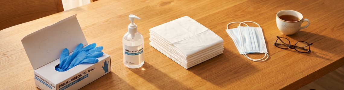 Pflegehilfsmittel zum Verbrauch: Handschuhe, Desinfektion und Bettschutz auf einem Holztisch