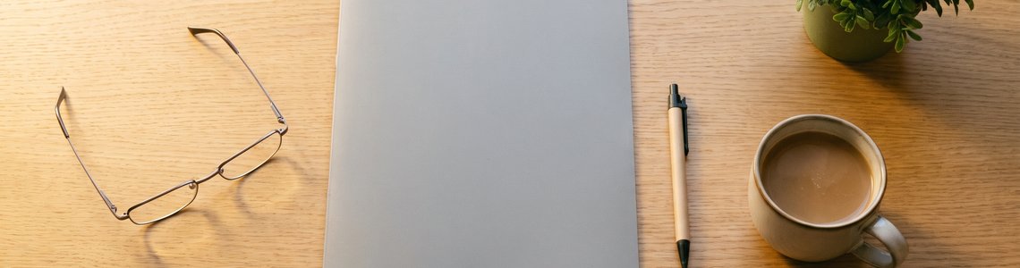 Schreibtisch mit Lesebrille, grauem Ordner, Kugelschreiber und Kaffeetasse als Symbol für die Planung des Rentenalters
