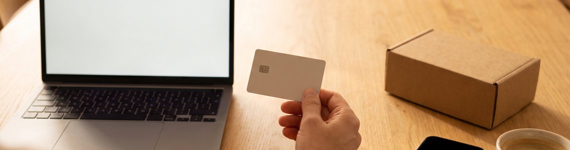 Hand mit Bankkarte vor einem Laptop am Küchentisch, daneben Smartphone und Paket