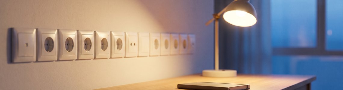 Steckdosen an einer Wohnzimmerwand und eine leuchtende Tischlampe im Abendlicht