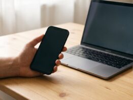 Hand mit Smartphone neben einem Laptop am Küchentisch als Sinnbild für den Login in App und Browser