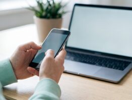 Hände halten ein Smartphone vor einem aufgeklappten Laptop bei der Anmeldung im Depot