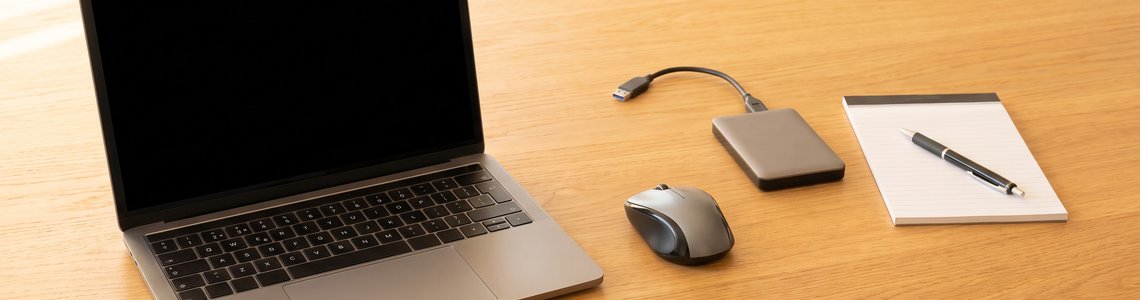 Aufgeklapptes Notebook mit dunklem Bildschirm, Maus, externer Festplatte und Notizblock auf einem Holzschreibtisch