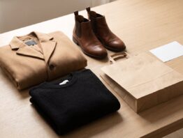 Gefalteter Wollmantel, schwarzer Strickpullover und Lederstiefeletten neben einer unbedruckten Papiertüte
